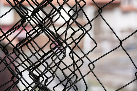 Bent chain link fence with distorted metal pattern. Close-up of damaged wire mesh with urban background. Abstract composition showing deformation and structure