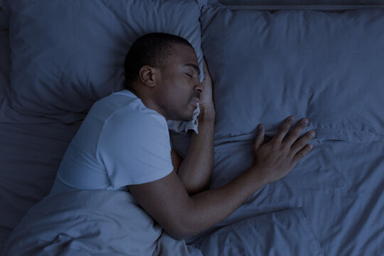 African american man sleeping at night in bed with blue tone lighting. Concept of deep sleep, nighttime rest and recovery in calm home environment, above view