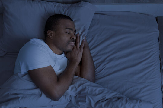 African american man sleeping at night in bed with hands under pillow. Concept of nighttime rest, recovery, calmness and deep sleep environment