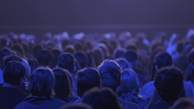 Large audience seated in dark theater under blue stage lighting, focused attention, shared anticipation, quiet atmosphere.