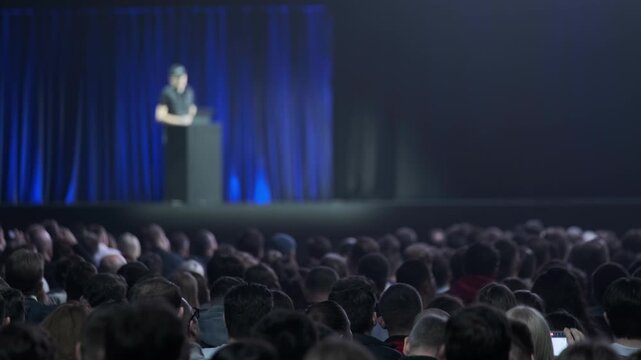 Large audience watches keynote speaker on stage. Corporate event atmosphere with blue curtain and spotlight. Focus on crowd and presentation energy.