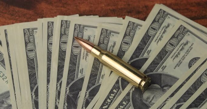 New shiny bullet set on top of a twenty dollar bills fan on a mahogany table symbolizing corruption and the costs of weapon trade now.