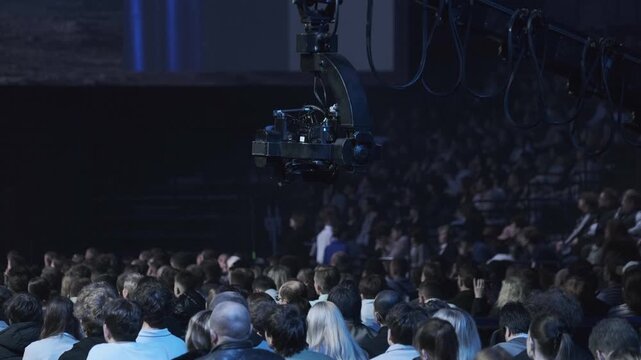 Large audience watching live concert while camera crane films stage, atmospheric blue lighting, professional production crew capturing performance.