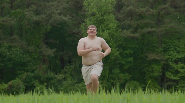 Fat man runs on grass. Outdoor fat jogging attempt. Fat body shows struggle. Fat loss motivation concept. Jogging man crosses green field. Beginner jogging shows effort. Overweight jogging.
