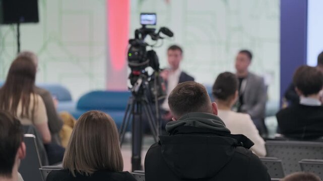 Audience listening to panel discussion at conference while camera records speakers, blurred foreground shows backs of attendees, corporate event atmosphere and media coverage.