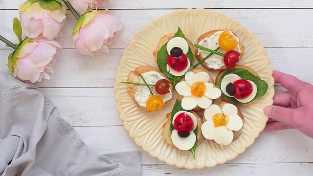 Hand placing a plate of spring or summer crostini on a white wood table. Summer garden party theme. Flowers, ladybugs.