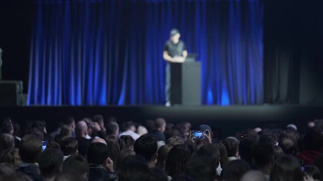 Large crowd watches speaker on stage at live conference event. Blue curtain backdrop, focused audience, phones recording, dim theater lighting.