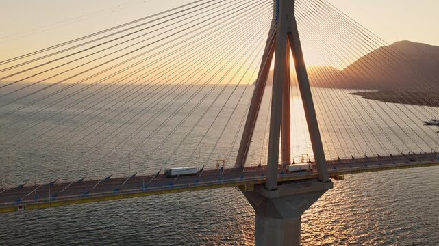 White cargo trucks travel across a scenic cable-stayed bridge at golden hour. A perfect aerial view of industrial transportation and global logistics at sunset.