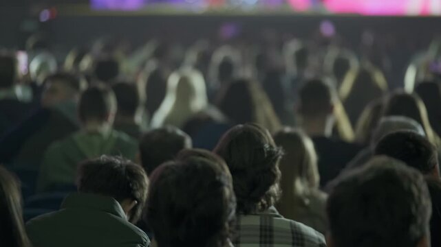 Large audience seated indoors facing stage, blurred shallow depth of field, mood of anticipation and focus, back of heads visible, event attendees watching presentation or performance.
