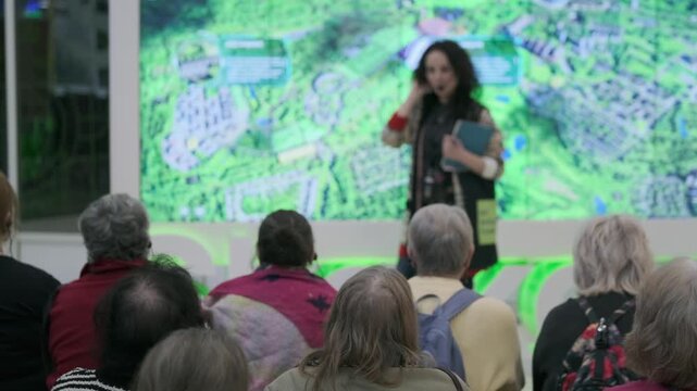 Senior audience attending community presentation about local development and city planning, presenter speaks in front of large green map display.