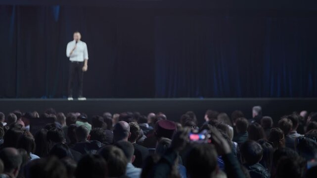 Large audience watches speaker on stage during corporate conference presentation, low lighting, blurred speaker, attendees record with smartphone.
