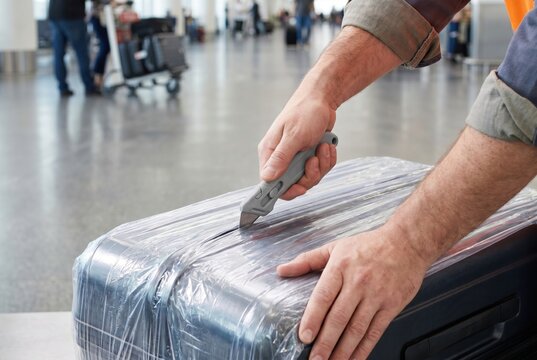 Airport worker hands using utility knife to cut plastic wrapping off a passenger suitcase