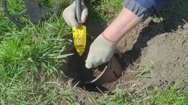 A man in gloves digs in the soil with a yellow trowel, discovering buried treasure in a hole, using a metal detector in a grassy open space