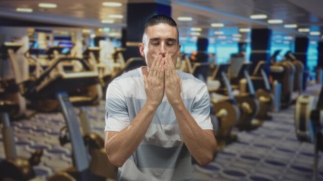 Man blows kiss hand to mouth in gym among treadmills and weight machines wearing striped t shirt; affection playful moment.