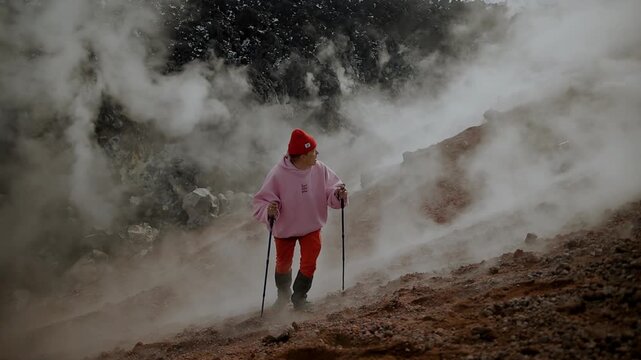 Young attractive man in red knitted hat and red pants goes up slope of volcano and looks back at solidified lava and clouds of smoke in crater. Concept of extreme recreation