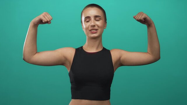 Woman flexing biceps with clenched fists against a green studio wall, short hair visible and toned arms on display; confidence empowerment.