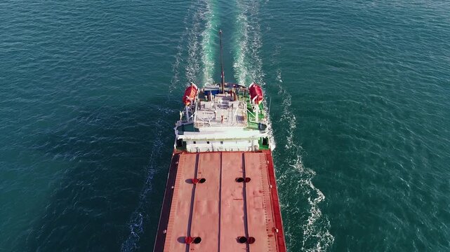 Aerial top-down view of dry cargo ship stern with water wake trailing behind on turquoise Black Sea, showing bridge deck equipment, lifeboats and red cargo hatch, Russia flag