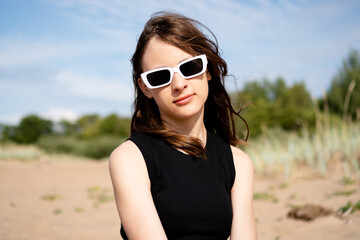 Naklejka premium A teenage girl poses on a sandy field with grass and trees behind, her calm gaze meeting the camera. The teenager radiates natural beauty and relaxed poise in this peaceful outdoor portrait.