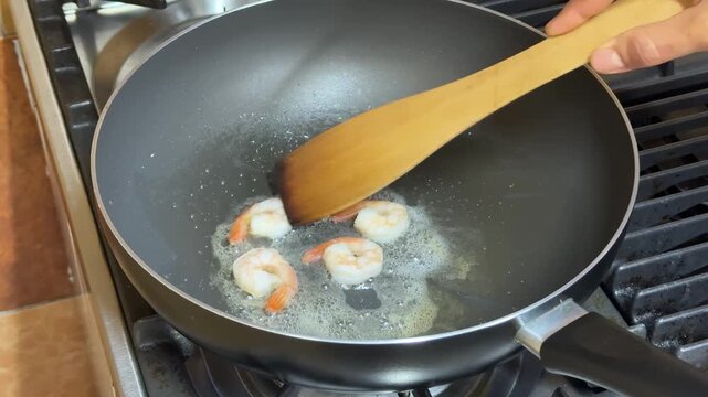 Hand stirs shrimp in nonstick skillet on gas stove, warm home kitchen scene, conveys everyday cooking and meal preparation
