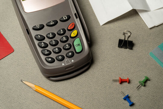 A card reader is on a gray table. Next to it are a pencil, colorful push pins, a binder clip, and some paper. This scene shows tools for work or transactions