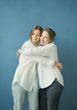 Beautiful mature mother and adult daughter in white shirts and jeans hugging in the photostudio on blue paper background. Two female generations. Mother's day concept