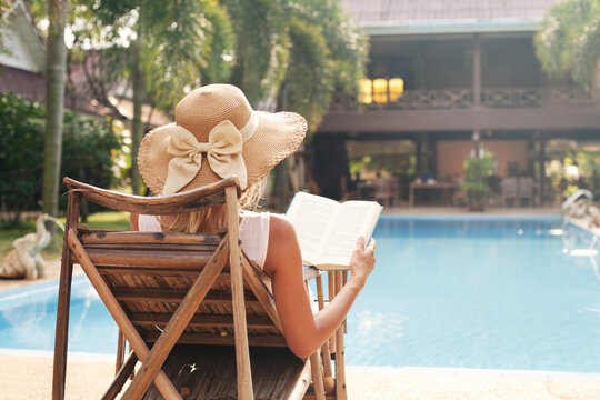 Unrecognizable woman in straw hat relaxing and reading book by hotel pool in tropical summer resort, vacation travel and relaxation.