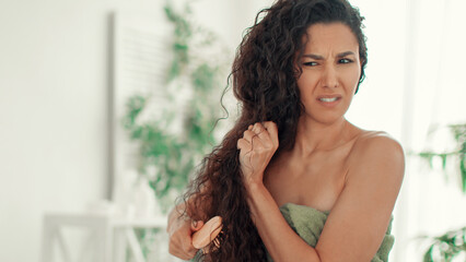 A woman stands in a well-lit room, brushing her curly hair. She is wrapped in a towel and looking at her hair with a confused expression. Green plants are visible in the background. © Prostock-studio