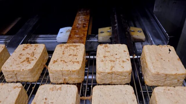 Smoked tofu blocks moving on a stainless steel wire mesh conveyor belt in a food processing factory. Industrial smoking production line.