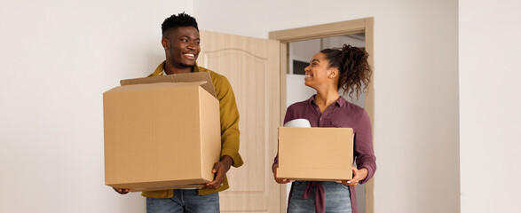 A couple stands in a light room carrying cardboard boxes. They smile at each other as they move into their new home. The setting shows a sense of excitement and teamwork during the moving process. © Prostock-studio