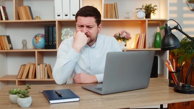 Young man on home table reads awful news on laptop and hits desk in anger from stress and frustration at home. Adult guy on home table reacts with shock and deep upset after bad online report indoors.