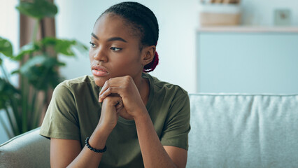 Fototapeta premium A young woman sits on a couch in a modern room. She has her chin resting on her hands and appears to be deep in thought. Green plants are visible in the background.