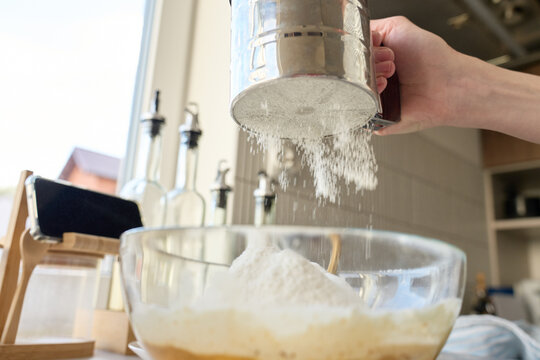 Hand sifting flour into glass bowl with batter on kitchen counter