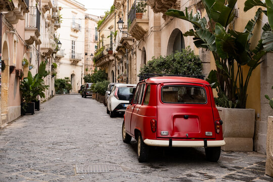 Vintage red car parked on a narrow cobblestone street in an old Italian town, surrounded by historic buildings and lush green plants