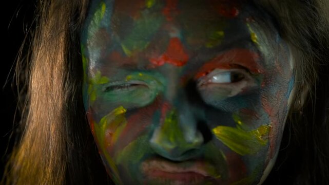 Woman with colorful face paint making funny expressions. Expressive woman with multicolored body art on her face making funny, strange, and silly faces while winking against a dark background