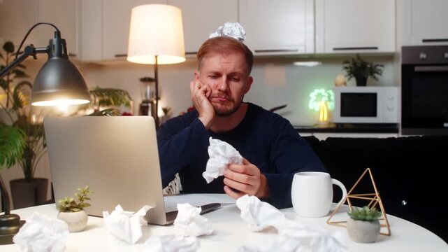 Young man at night home works on project writes ideas fights creative block feels burnout mood. Caucasian guy on sofa at table stares at laptop sees crumpled papers critiques plans next step steady.
