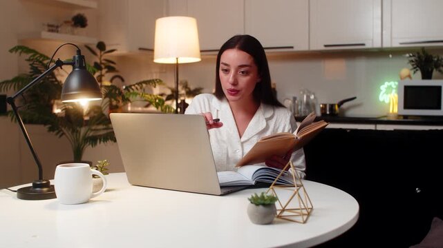 Young woman at night home studies on laptop watches tutorial reads book takes notes solves quiz. Caucasian girl on sofa at evening reviews topics writes key points prepares exam stays calm focus well.