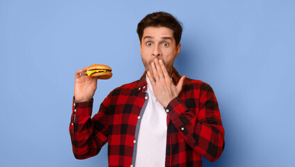 A man stands with a hamburger in one hand and his other hand covering his mouth. He looks surprised while wearing a checkered shirt against a blue background. © Prostock-studio