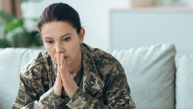 A woman wearing a military uniform sits on a couch with a thoughtful expression. She has her hands clasped together, reflecting on her service and experiences.