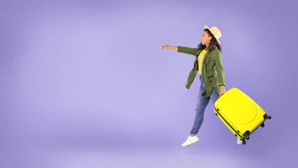 A woman stands on a purple background holding a yellow suitcase. She is wearing casual clothes and smiles while pointing ahead, as if excited about her travel plans. © Prostock-studio