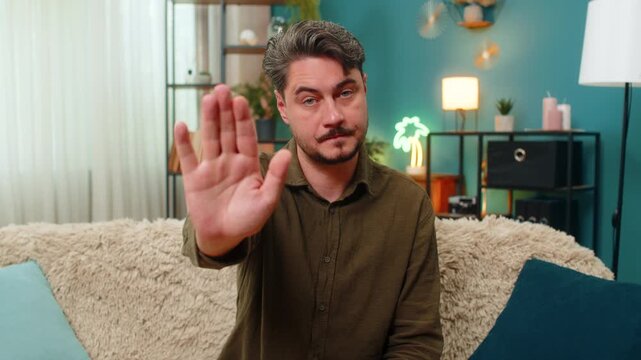 Middle-aged man at home raises hand making stop gesture holding stern gaze rejecting action. Young adult guy on sofa signals halt shows caution strict boundary negative response unwilling continue no