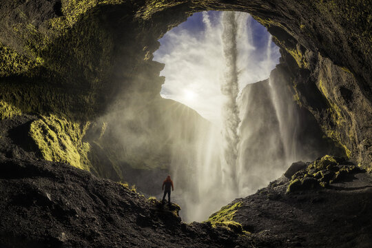 View of Kvernufoss waterfall from inside a mossy cave with a person in a red jacket standing on a ledge amidst mist and sunlight in Evindarholar, Rangarthing eystra, Iceland.