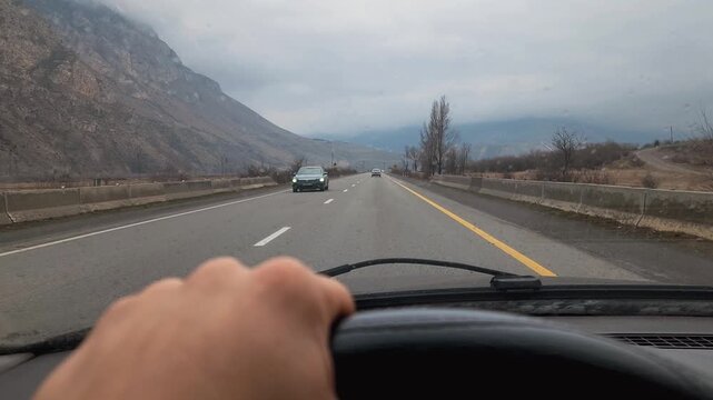 Driver's point of view traveling on a two-lane mountain highway on an overcast day, with a hand visible on the steering wheel and other cars in the distance moving through the landscape