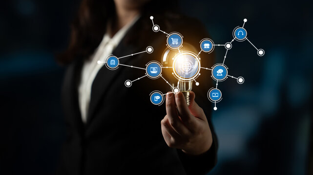 A focused businesswoman presents a lightbulb amidst digital icons, symbolizing innovation, technology, and networking. Ideal for themes of creativity and progress in a corporate setting. Tessel