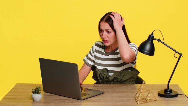 Young woman sitting at table aggressively typing and suddenly slamming laptop closed in frustration with system failure. Girl on yellow background shows anger and emotional reaction to technical error