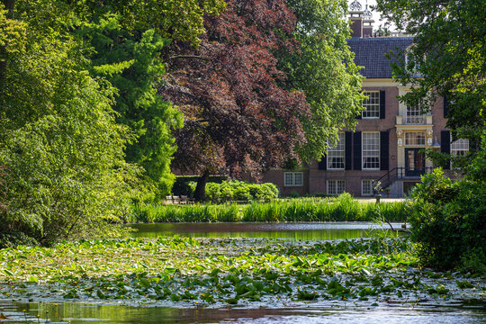 View of Castle Groeneveld with its historic brick facade framed by lush green trees and a pond filled with lily pads in Baarn, Utrecht, Netherlands.