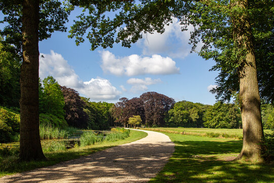 View of a winding gravel path through the lush estate parkland of Castle Groeneveld with mature trees and a pond in Baarn, Utrecht, Netherlands.