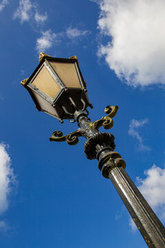 View of an ornate vintage street lamp with gold decorative elements against a bright blue sky with white clouds at Castle Groeneveld in Baarn, Utrecht, Netherlands.