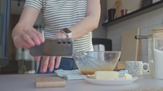 Woman whisking batter in glass bowl while holding smartphone in kitchen. Concept of dessert preparation and online recipe use
