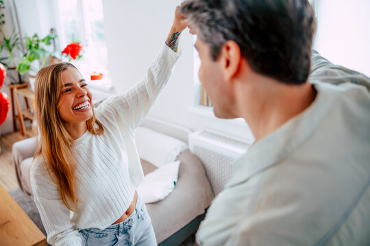 Smiling young woman raises her arm while dancing with her partner in a cozy living room. Happy couple enjoying a playful, romantic moment at home filled with light, warmth, and love.