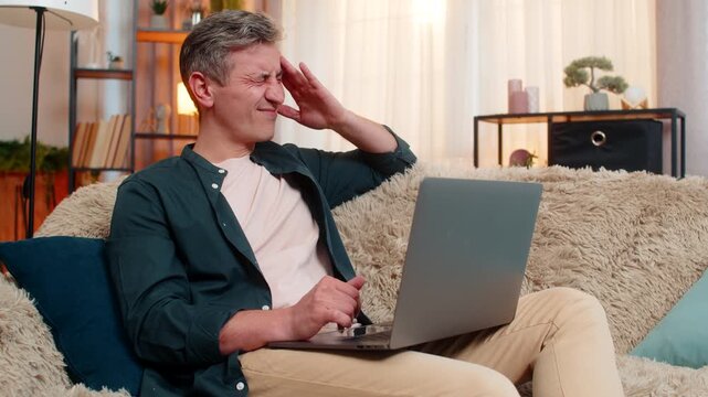 Young man at home looks exhausted while holding head and massaging temples in front of laptop. Caucasian guy on sofa appears tired and overworked after long hours of concentration and mental effort.